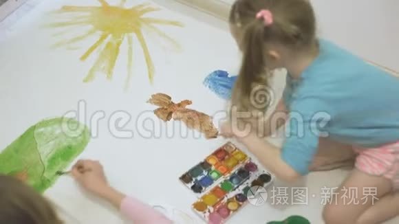 两个女孩在地板上用水彩颜料和画笔在一张大画纸上画画，孩子们`共同的创造力。
