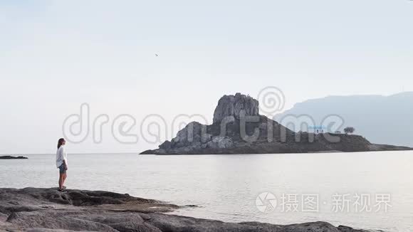 一个年轻的女人正在小岛的背景下走在岩石海岸上。