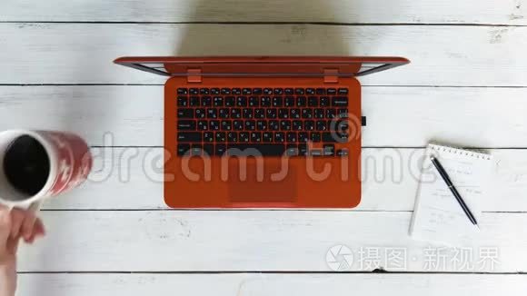 一个男人在一台红色的笔记本电脑上打字，笔记本电脑放在一张白色的木桌上。左边是一杯黑咖啡