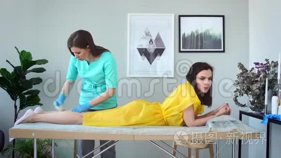 两个女孩在一家美容院，主人穿着浴衣，戴着手套，做着用糖去除腿上毛发的程序
