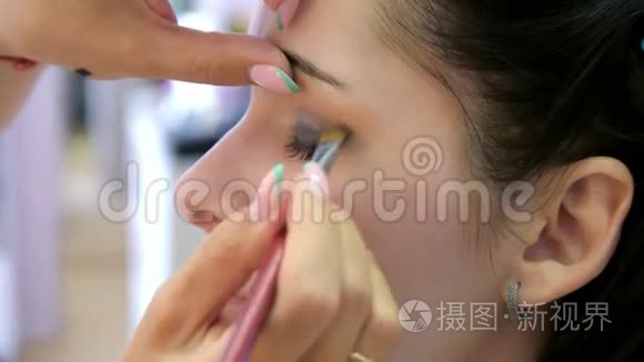 双手化妆师将化妆涂在一个年轻女人的脸上。 在专业沙龙里做眼妆的女孩。 晚上