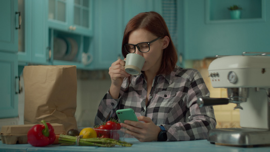 戴眼镜的年轻成年女性喝咖啡用手机站在家里的蓝色厨房视频