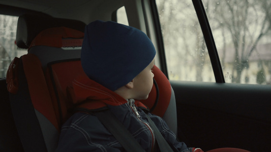 男孩坐在儿童座位上的汽车中骑着男孩在雨季看着窗外视频