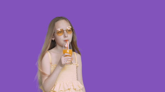 少女在紫罗兰色背景上用稻草从玻璃杯中喝橙汁戴着黄色视频