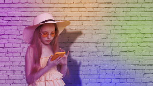 十几岁的女孩在五颜六色的灯光下在砖墙背景上浏览手机视频