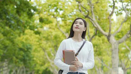 年轻女孩抱着书本走在校园林荫道视频