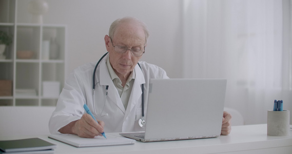 老年医生在网上咨询看笔记本电脑屏幕和纸面写笔记时倾听视频
