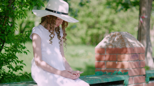 卷曲头发的女孩在夏天花园里使用智能手机在长凳上穿着白帽子视频