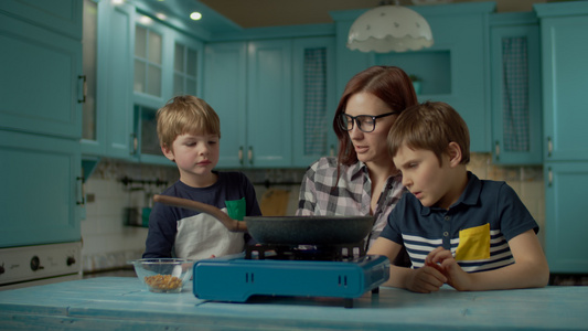 年轻母亲和两个儿子在家里蓝色厨房的煎锅上煮爆米花的视频