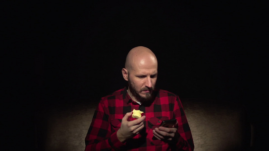 有胡子吃苹果的年轻的青年秃头长发男子在智能手机视频