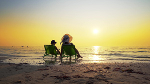 母亲和儿子戴着帽子坐在沙滩椅上看着海浪溅起的落日旅游24秒视频