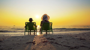 母亲和儿子戴着帽子坐在沙滩椅上看着海浪和落日17秒视频