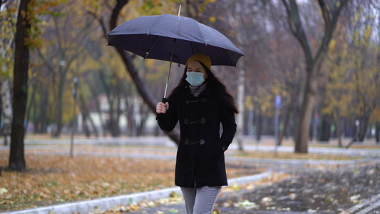 一位戴着防护面具的年轻女子在雨伞下在公园里散步下雨天视频