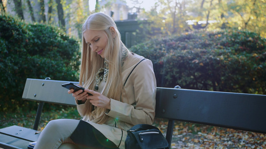 金发女孩在公园户外的长凳上用手机视频
