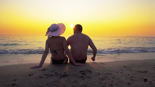 一对浪漫的情侣戴着帽子的男人和女人坐在海边看着日落视频