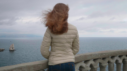 年轻苗条的女人长着棕色的长发在刮风的日子里站在阳台视频