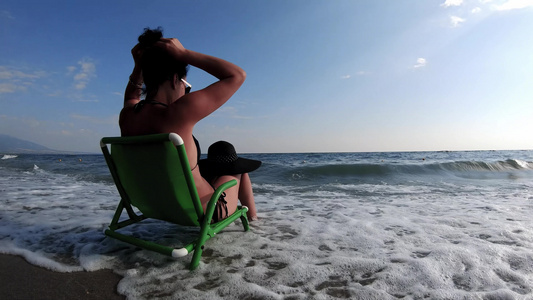 穿着比基尼的漂亮年轻美女戴着帽子坐在沙滩上的椅子上视频