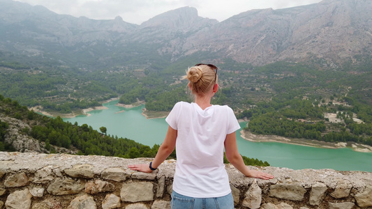 一位年轻女子看着一个美丽的风景蓝湖和山峰从一座古老视频