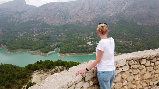 一位年轻女子看着一个美丽的风景蓝湖和山峰从一座古老视频