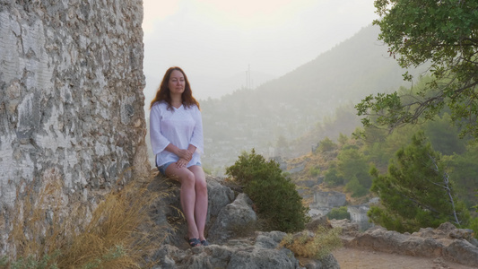 年轻女子坐在悬崖背景上的石头上旅游女人在青山背景上视频