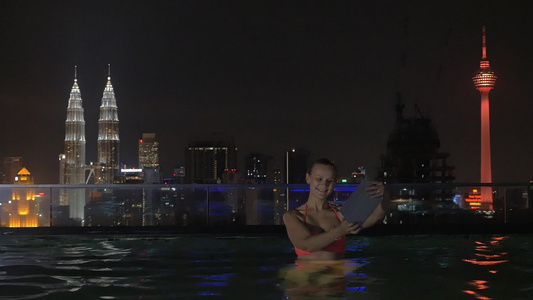 在摩天大楼屋顶上游泳池中女性用平板制作自拍与夜间城市视频