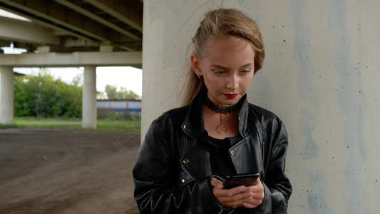 穿着黑色皮夹克的年轻女孩在城市街道上使用手机视频