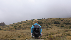 穿着蓝色夹克的女性徒步旅行者背着背包坐在岩石上欣赏10秒视频