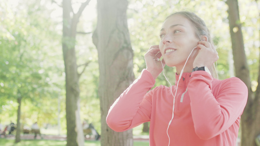 穿着粉红运动服的健身女孩在公园跑时用耳机听音乐和听视频