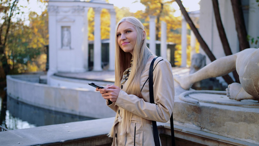 在秋天公园穿着米花大衣的金发美女看她的智能手机视频