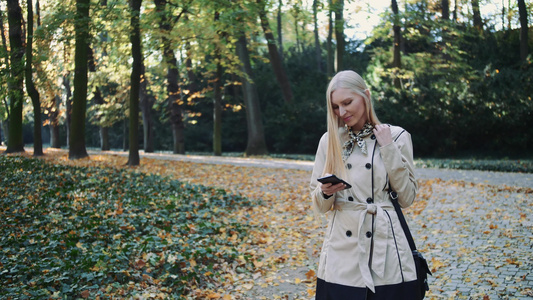 在秋天公园散步看着智能手机和打字的金发女子视频