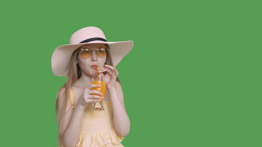女孩少年用吸管从透明绿色背景的玻璃中喝橙汁戴着黄色视频