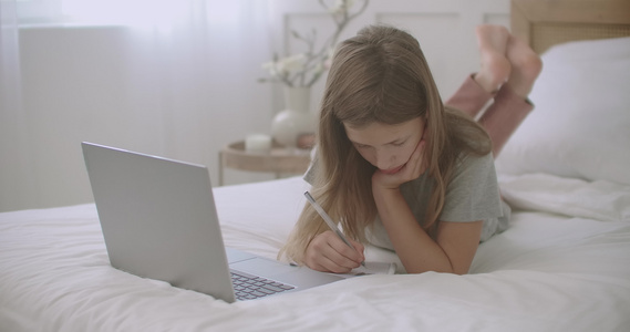 无聊的小女孩正在抄写本上做作业在网上上课时看笔记本电脑视频
