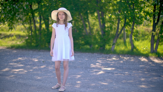 穿着白色连衣裙和帽子的年轻女孩站在绿树背景的公园小巷视频