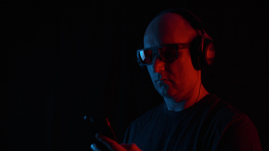 成年男子用耳机在黑色背景上用红色和蓝色灯光听移动音乐视频