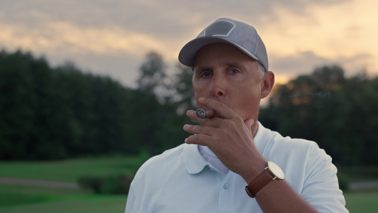 高级高尔夫球手在日落球场球道上抽着雪茄看着相机视频