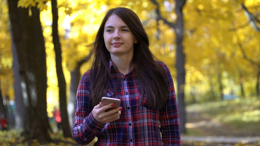 秋天女孩在公园散步在她的智能手机上使用应用程序录像片视频