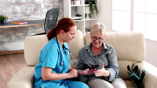 坐在沙发上帮助年长妇女使用智能手机的疗养院护理员视频