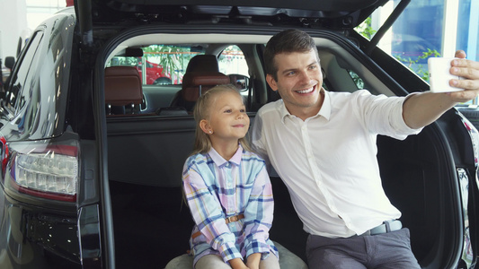 年轻父亲和女儿在汽车背景上做自拍并用车背面制作自拍视频