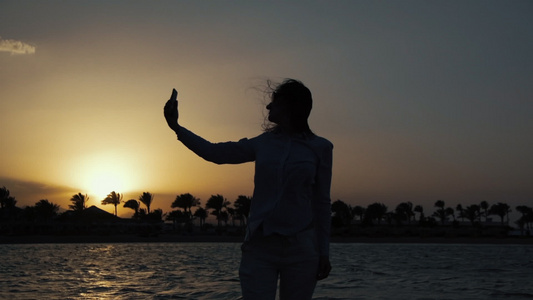 美丽女性在日落海边上用手机自拍逆光背影视频