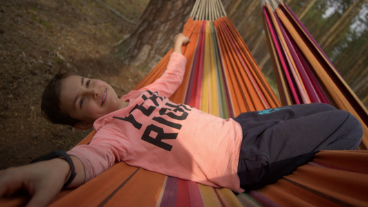 小男孩快乐地面对镜头躺在秋天树林的吊床上享受时光视频