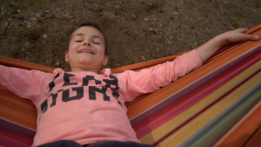 小男孩快乐地面对镜头躺在秋天树林吊床上享受时光正面特写视频