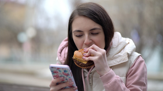 快乐的女人吃汉堡在街上用智能手机视频