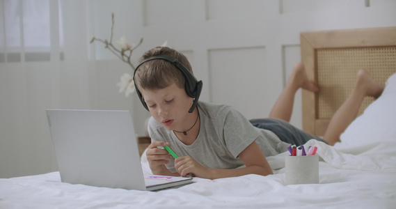 青少年男孩在家里床上的专辑上画彩色和图片用笔记本电脑视频