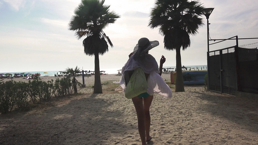 穿着帽子和围巾的性感女人在棕榈树之间的沙滩上走下视频