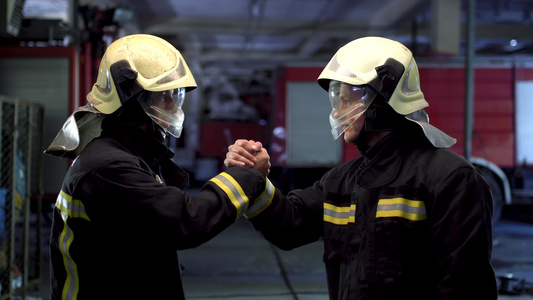 两名身着制服和防护头盔的消防员握手致意消防车在背景视频