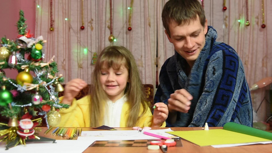 爸爸坐在她女儿旁边笑着扔了一瓶胶水来帮她做圣诞手工艺品视频