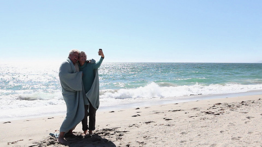 一对成熟的夫妇在沙滩上拿着毯子使用智能手机给自己拍照视频