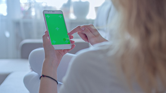 身穿白色牛仔裤的年轻妇女躺在沙发上用绿色屏幕使用智能手机视频