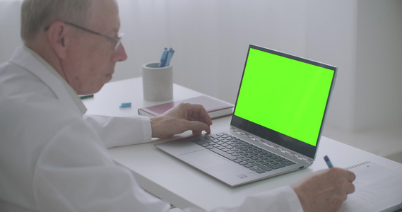 老年男性医生正在互联网上阅读文章查看笔记本电脑和写作视频