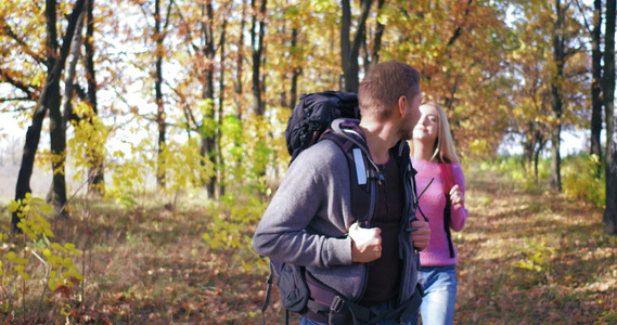 在阳光明媚的下午情侣穿过秋天的森林背着背包看着两边视频